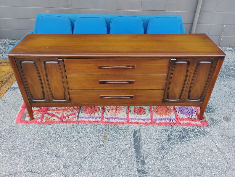 May include: A long wooden sideboard featuring a smooth rectangular top, three central horizontal drawers with dark wood handles, and two side cabinets with recessed panel doors. The piece stands on four tapered legs and has a warm brown finish.
