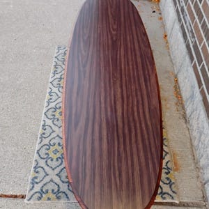 May include: Oval-shaped wooden coffee table with a dark brown wood grain finish. The table has a lighter brown trim around the edge. The table is approximately 60 inches long and 24 inches wide. The table is on a patterned rug.