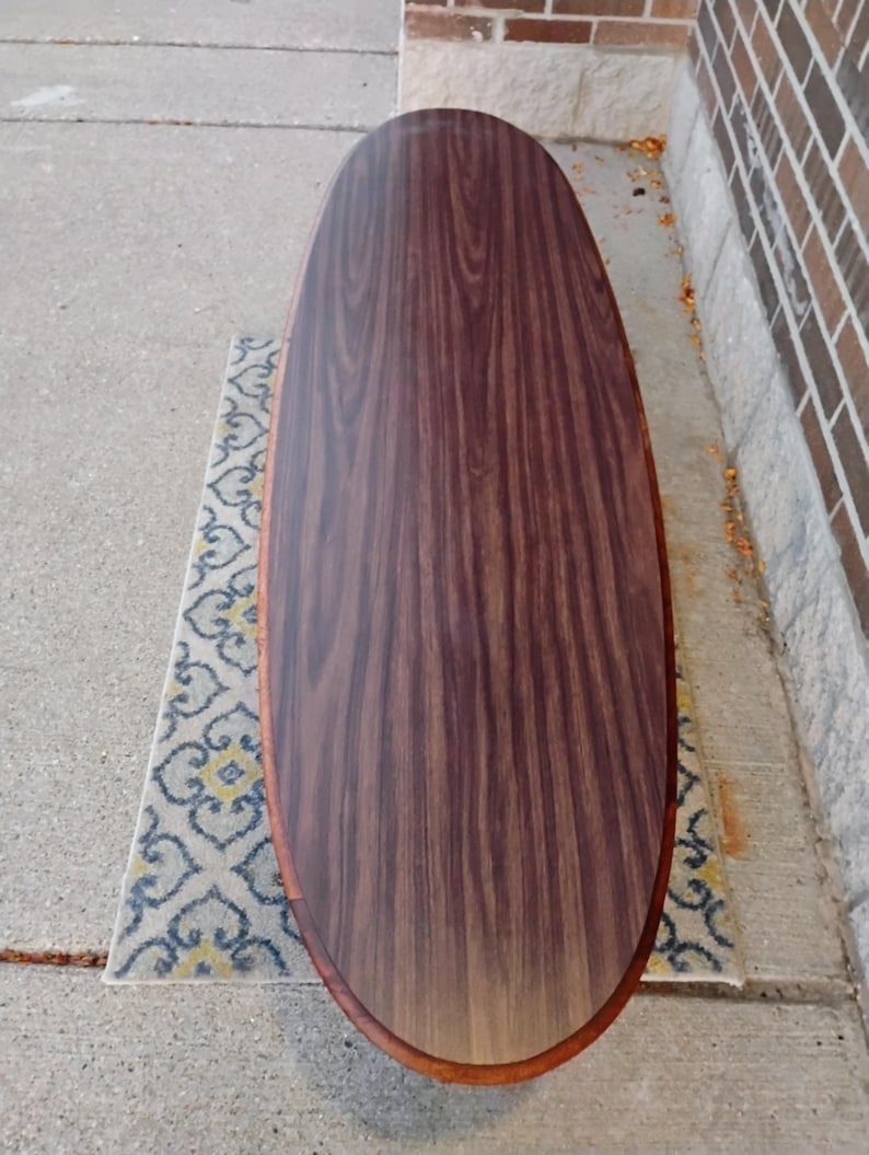 May include: Oval-shaped wooden coffee table with a dark brown wood grain finish. The table has a lighter brown trim around the edge. The table is approximately 60 inches long and 24 inches wide. The table is on a patterned rug.
