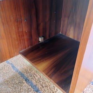 May include: An open wooden cabinet interior featuring a rich, dark brown wood grain finish with visible vertical patterns. The cabinet includes a single horizontal shelf and a small, light-colored square block attached to the back panel interior.