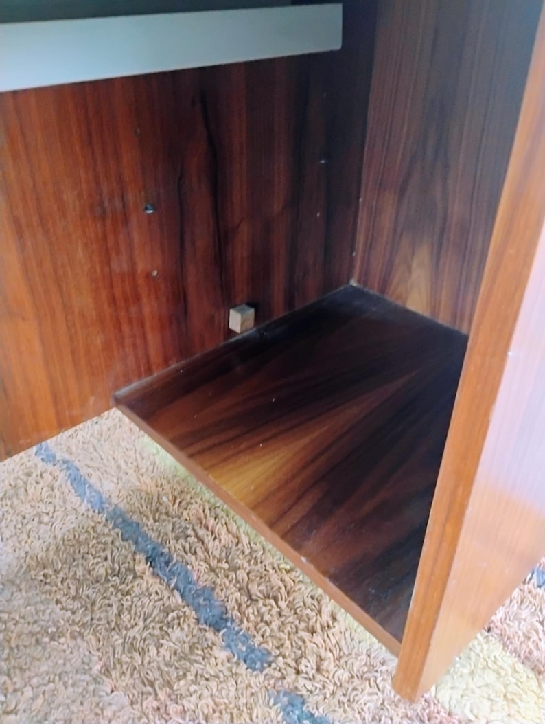 May include: An open wooden cabinet interior featuring a rich, dark brown wood grain finish with visible vertical patterns. The cabinet includes a single horizontal shelf and a small, light-colored square block attached to the back panel interior.