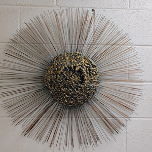 Puede incluir: Una gran escultura de pared redonda de color dorado con un centro texturizado y p&uacute;as met&aacute;licas radiantes.