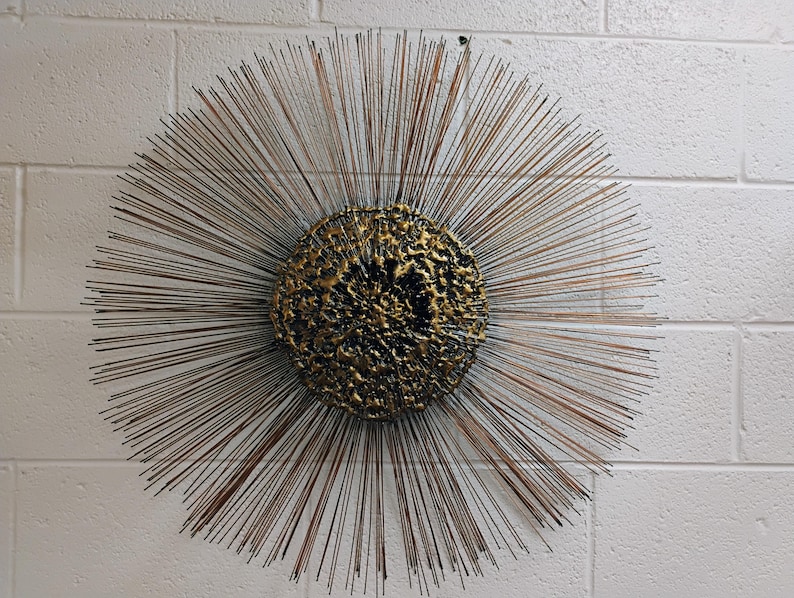 Puede incluir: Una gran escultura de pared redonda de color dorado con un centro texturizado y p&uacute;as met&aacute;licas radiantes.