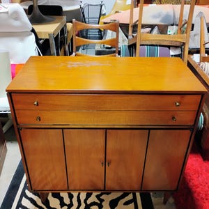Puede incluir: Un armario de madera de estilo moderno de mediados de siglo con dos cajones y dos puertas. El armario est&aacute; hecho de madera marr&oacute;n claro con un acabado natural. El armario tiene herrajes de lat&oacute;n.