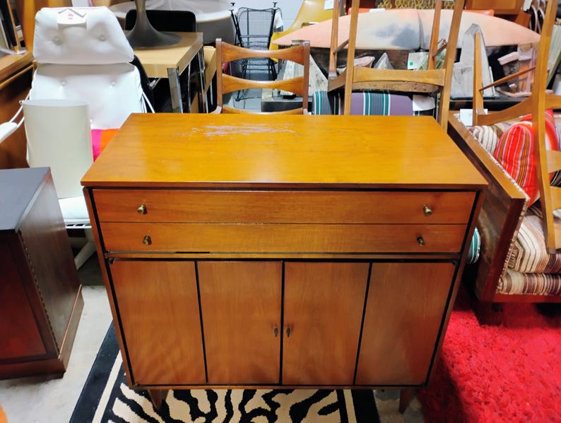 Puede incluir: Un armario de madera de estilo moderno de mediados de siglo con dos cajones y dos puertas. El armario est&aacute; hecho de madera marr&oacute;n claro con un acabado natural. El armario tiene herrajes de lat&oacute;n.