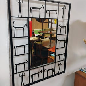 Puede incluir: Un espejo con marco de metal negro con un diseño geométrico que presenta figuras estilizadas de personas y animales. El espejo refleja una habitación con muebles y otros objetos.
