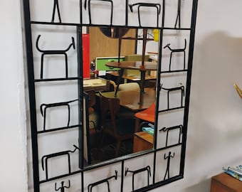 May include: A black metal framed mirror with a geometric design featuring stylized figures of people and animals. The mirror reflects a room with furniture and other objects.
