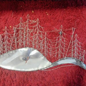 Pode incluir: Uma pe&ccedil;a de arte de parede em metal que retrata uma paisagem de inverno. A obra apresenta &aacute;rvores prateadas estilizadas e uma base prateada curva e reflexiva, possivelmente representando uma colina ou lago coberto de neve. O fundo &eacute; vermelho escuro.