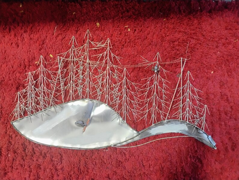 Pode incluir: Uma pe&ccedil;a de arte de parede em metal que retrata uma paisagem de inverno. A obra apresenta &aacute;rvores prateadas estilizadas e uma base prateada curva e reflexiva, possivelmente representando uma colina ou lago coberto de neve. O fundo &eacute; vermelho escuro.