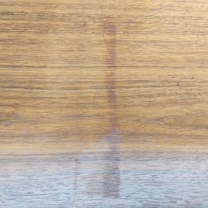 May include: A flat wooden surface featuring a prominent horizontal grain pattern in shades of light brown and tan. A vertical, darker brown discoloration or stain runs down the centre of the wood, contrasting with the lighter natural wood tones.