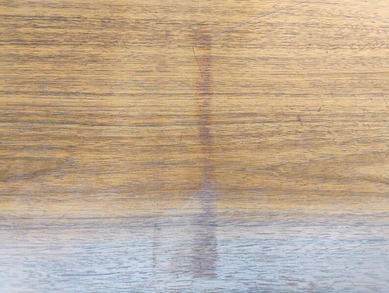 May include: A flat wooden surface featuring a prominent horizontal grain pattern in shades of light brown and tan. A vertical, darker brown discoloration or stain runs down the centre of the wood, contrasting with the lighter natural wood tones.