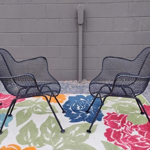 May include: Four black metal mesh chairs with a floral patterned rug underneath. The chairs have a curved back and are made of a wire mesh material.