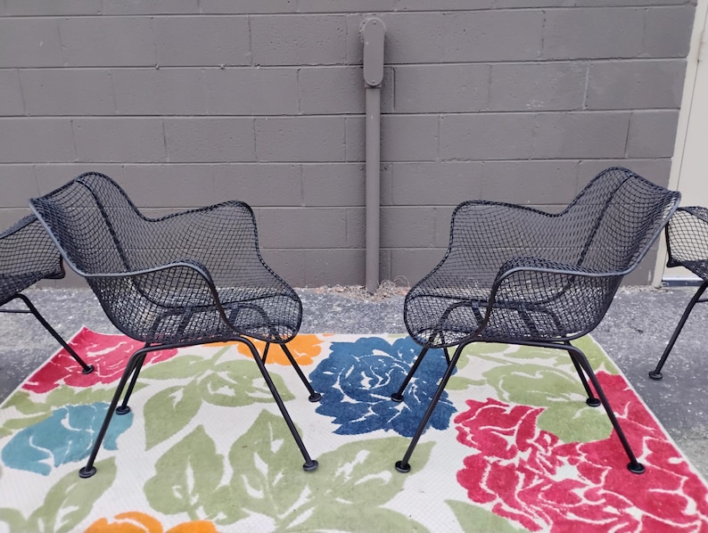 May include: Four black metal mesh chairs with a floral patterned rug underneath. The chairs have a curved back and are made of a wire mesh material.