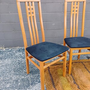 May include: Two high-back chairs constructed from light-toned wood, each featuring a square lattice design in the upper backrest. The seats are upholstered in a solid black fabric, and the chairs have four straight legs with connecting supports.