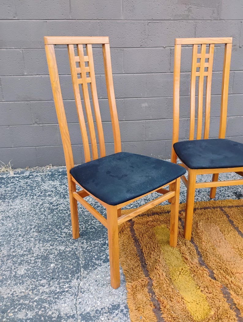 May include: Two high-back chairs constructed from light-toned wood, each featuring a square lattice design in the upper backrest. The seats are upholstered in a solid black fabric, and the chairs have four straight legs with connecting supports.
