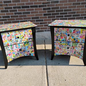 May include: Two black nightstands with comic strip collage fronts and tops. Each nightstand has two drawers and curved legs. The comic strip collage features various colorful cartoon characters and scenes.