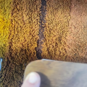May include: A shaggy rug featuring vertical stripes in shades of yellow, mustard, and dark brown. The texture is dense and looped, creating a thick pile surface. The rug displays a gradient color pattern with distinct color transitions between the stripes.