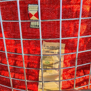 May include: Close-up of a vintage chair with a red woven seat and a chrome metal grid back. The seat has a textured pattern. A label with text is visible on the back of the chair.