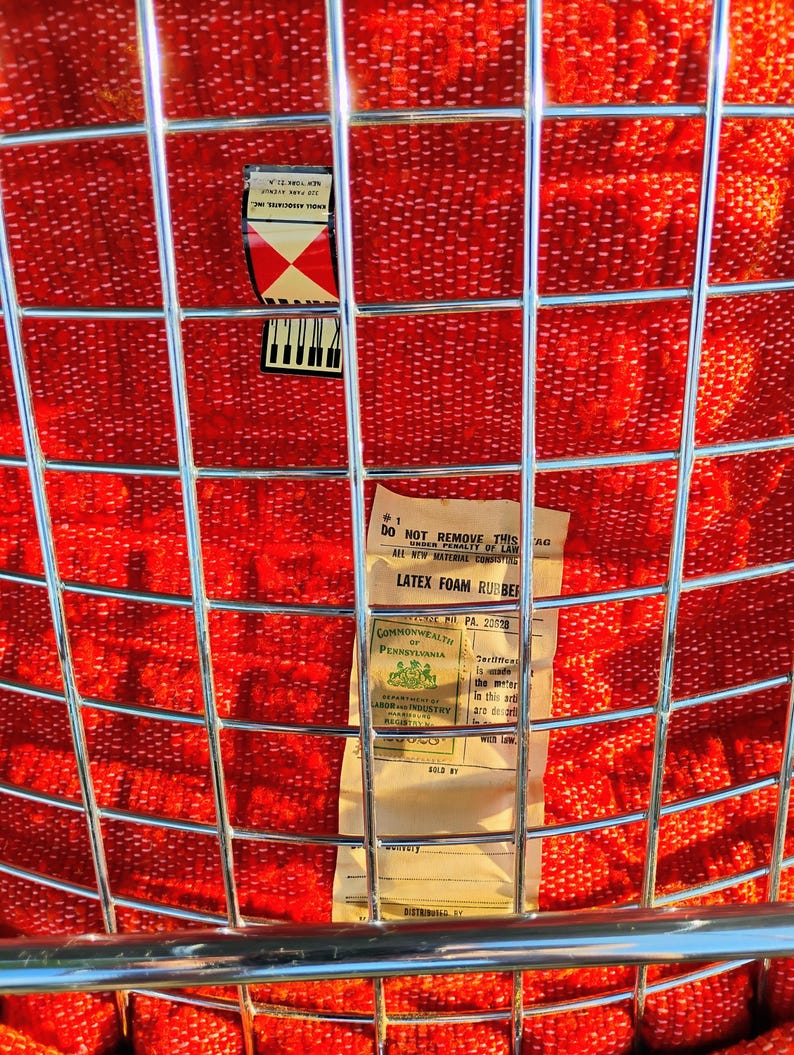 May include: Close-up of a vintage chair with a red woven seat and a chrome metal grid back. The seat has a textured pattern. A label with text is visible on the back of the chair.