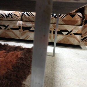 May include: A close-up view of a mid-century modern sofa with a geometric patterned fabric in shades of brown, black, and cream. The sofa has a dark wooden frame and metal legs. A shaggy brown rug is visible in the foreground.