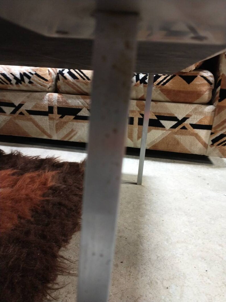 May include: A close-up view of a mid-century modern sofa with a geometric patterned fabric in shades of brown, black, and cream. The sofa has a dark wooden frame and metal legs. A shaggy brown rug is visible in the foreground.