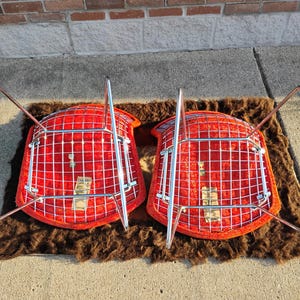 May include: Two mid-century modern chairs with bright red seats and chrome legs are displayed upside down on a brown rug. The chairs have a grid-like design on the seat backs.