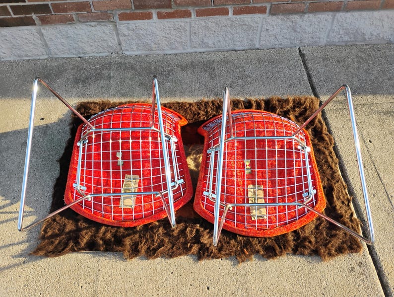 May include: Two mid-century modern chairs with bright red seats and chrome legs are displayed upside down on a brown rug. The chairs have a grid-like design on the seat backs.