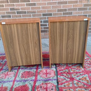 May include: Two wooden cabinets with a light brown finish and a wood grain pattern. The cabinets are rectangular with a darker brown top and are placed on a colorful rug. The cabinets are against a brick wall.