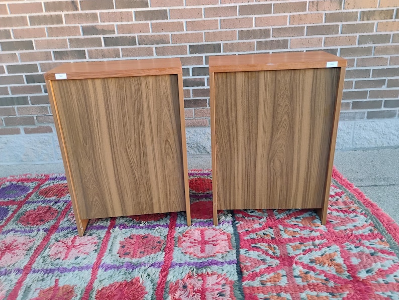 May include: Two wooden cabinets with a light brown finish and a wood grain pattern. The cabinets are rectangular with a darker brown top and are placed on a colorful rug. The cabinets are against a brick wall.
