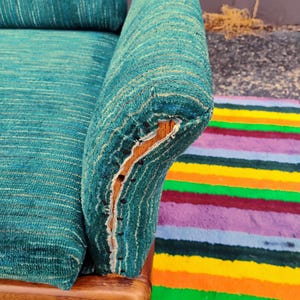 May include: A teal textured fabric sofa armrest with a visible wooden frame and exposed upholstery tacks along the side. The fabric features a subtle horizontal weave pattern in shades of blue and green, with a worn edge revealing the wood underneath.
