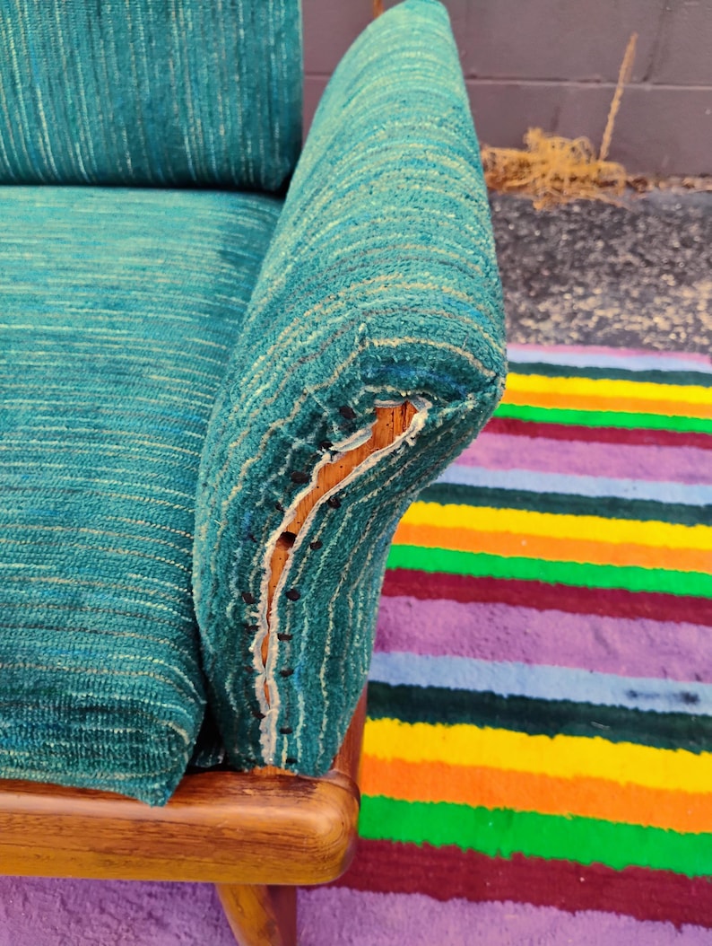 May include: A teal textured fabric sofa armrest with a visible wooden frame and exposed upholstery tacks along the side. The fabric features a subtle horizontal weave pattern in shades of blue and green, with a worn edge revealing the wood underneath.