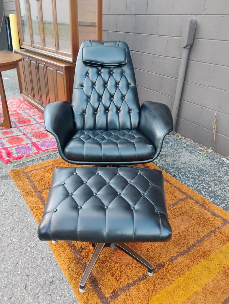 May include: A black leather lounge chair and matching ottoman set featuring a tufted button design, a high backrest with an integrated headrest, curved armrests, and a metal swivel base. The upholstery has a smooth finish with diamond patterns.