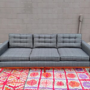 K&ouml;nnte beinhalten: Ein Dreisitzer-Sofa, bezogen mit einem dunkelgrauen Stoff mit dezentem Karomuster. Das Sofa verf&uuml;gt &uuml;ber drei gesteppte R&uuml;ckenkissen, drei Sitzkissen und einen h&ouml;lzernen Unterbau mit vier Beinen in einem klaren, linearen Design.