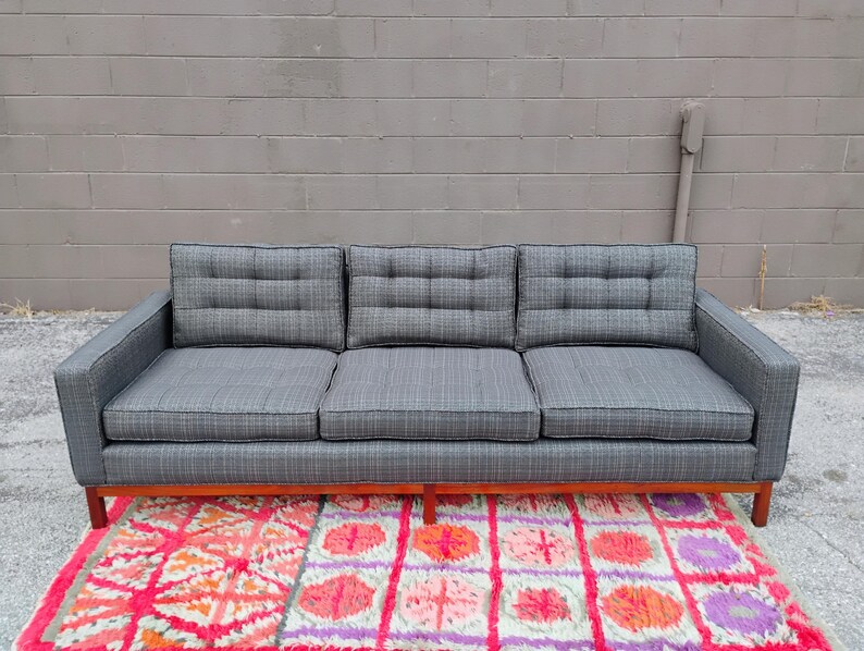 K&ouml;nnte beinhalten: Ein Dreisitzer-Sofa, bezogen mit einem dunkelgrauen Stoff mit dezentem Karomuster. Das Sofa verf&uuml;gt &uuml;ber drei gesteppte R&uuml;ckenkissen, drei Sitzkissen und einen h&ouml;lzernen Unterbau mit vier Beinen in einem klaren, linearen Design.