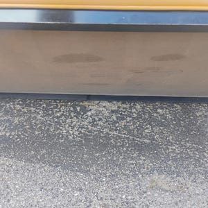 May include: A horizontal rectangular object featuring a smooth, light brown surface bordered by a thin black frame. Above the black frame, a narrow strip of yellow material is visible. The object is positioned against a dark, textured asphalt surface.