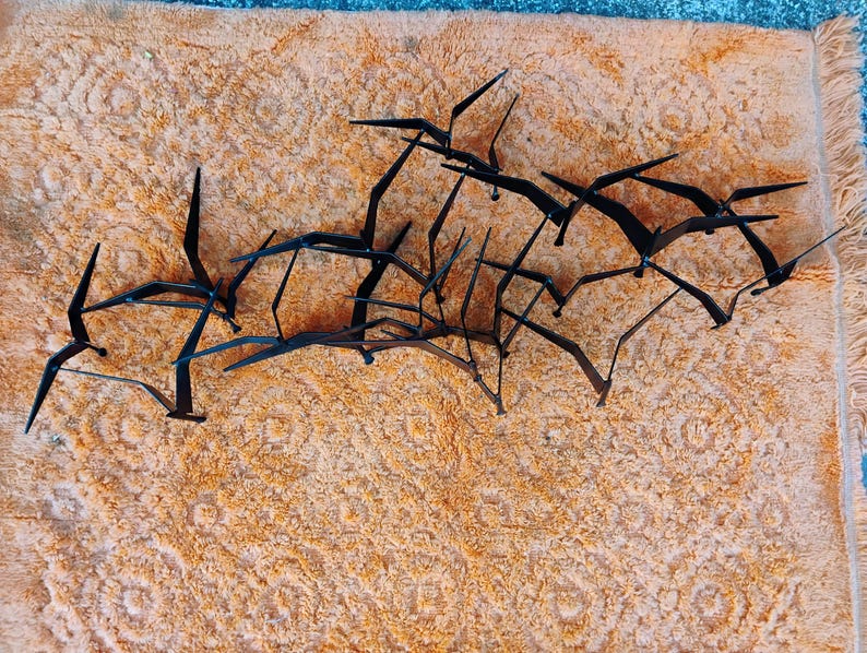 May include: Black metal bird sculpture with multiple birds in flight. The sculpture is set against a textured, orange background. The birds have outstretched wings, creating a dynamic and artistic display.