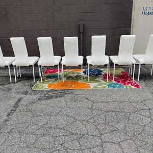 May include: Six white dining chairs with a sleek design and metal legs are arranged in a row. They are positioned on a colorful floral patterned rug with orange, red, blue, and green hues.