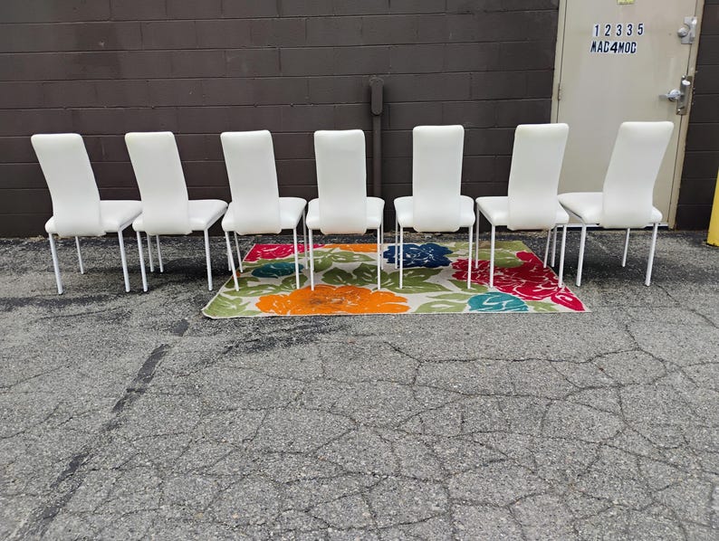 May include: Six white dining chairs with a sleek design and metal legs are arranged in a row. They are positioned on a colorful floral patterned rug with orange, red, blue, and green hues.