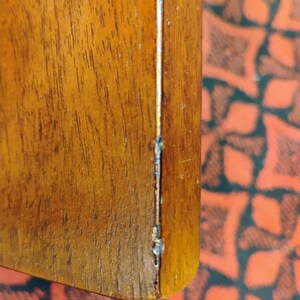 May include: A close-up view of a smooth, medium-brown wooden surface with a visible vertical grain pattern. Along the right edge, there is a distinct, narrow seam or joint line that shows signs of wear, including minor chipping and dark discoloration.