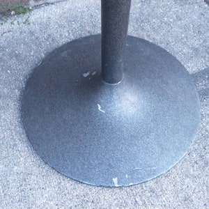 May include: Close-up of a gray metal table base with a circular top and a single, central support post. The base has a slightly textured surface.