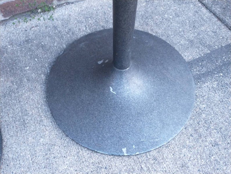 May include: Close-up of a gray metal table base with a circular top and a single, central support post. The base has a slightly textured surface.