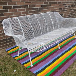 May include: A white metal outdoor bench featuring a grid-patterned mesh seat and backrest. The bench has a curved silhouette with integrated armrests and is supported by four thin, angled metal legs that provide a stable base for the structure.