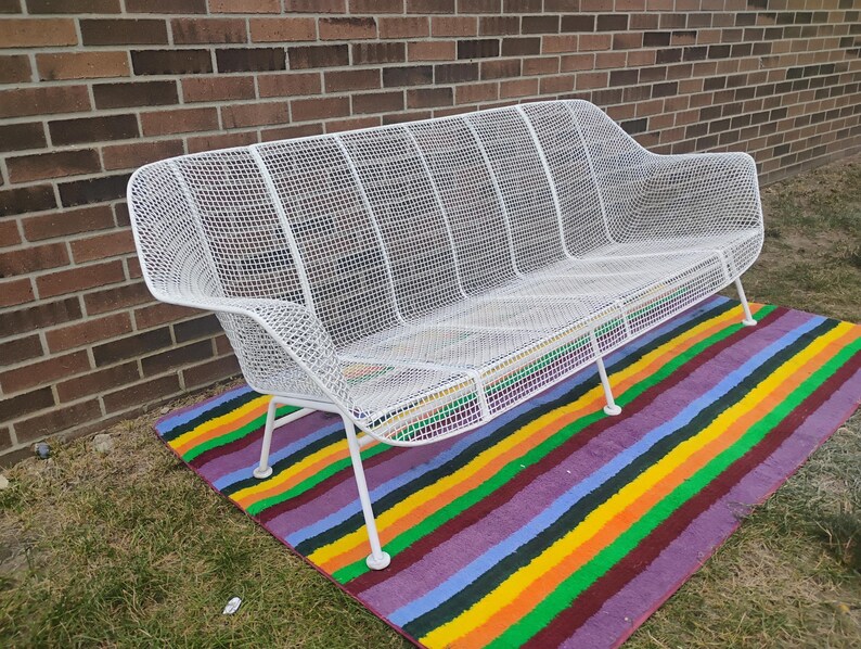 May include: A white metal outdoor bench featuring a grid-patterned mesh seat and backrest. The bench has a curved silhouette with integrated armrests and is supported by four thin, angled metal legs that provide a stable base for the structure.