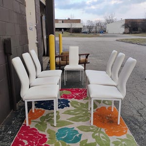 May include: A set of six white upholstered dining chairs with a curved back and a white metal frame. The chairs are arranged around a colorful floral rug with red, orange, blue, and green flowers. A single white chair is also visible.
