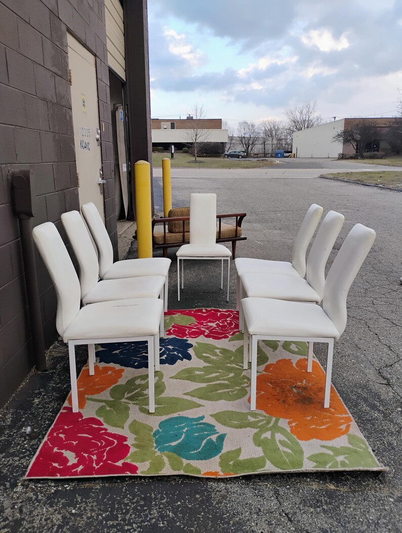 May include: A set of six white upholstered dining chairs with a curved back and a white metal frame. The chairs are arranged around a colorful floral rug with red, orange, blue, and green flowers. A single white chair is also visible.