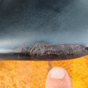 May include: A black, smooth, curved surface featuring a prominent, textured, dark brown scuff mark along the edge. The material appears to have a slight sheen, and the damaged area shows a rough, worn-down texture contrasting with the surrounding finish.