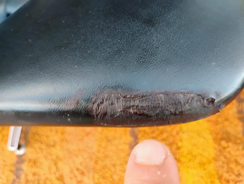 May include: A black, smooth, curved surface featuring a prominent, textured, dark brown scuff mark along the edge. The material appears to have a slight sheen, and the damaged area shows a rough, worn-down texture contrasting with the surrounding finish.