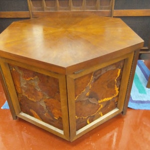 May include: A mid-century modern hexagonal cabinet with a wooden top and sides. The cabinet doors feature a unique, abstract design in shades of brown, orange, and yellow. The cabinet is approximately 30 inches wide and 20 inches tall.