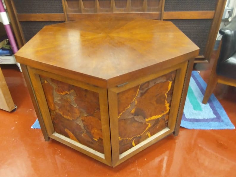 May include: A mid-century modern hexagonal cabinet with a wooden top and sides. The cabinet doors feature a unique, abstract design in shades of brown, orange, and yellow. The cabinet is approximately 30 inches wide and 20 inches tall.