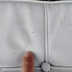 May include: Close-up of a white leather surface with visible wear and tear, including small dark spots and scratches. The surface is divided into sections by stitched seams and a button is visible.